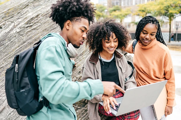 Three people are gathered outdoors, smiling and looking at a laptop.
